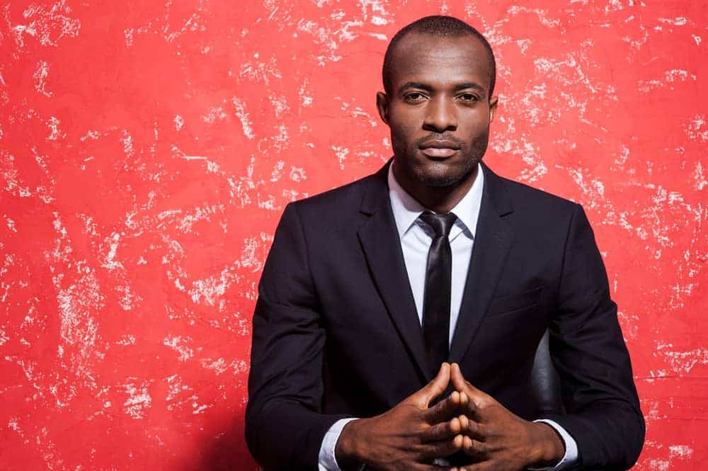 Hombre de negocios afroamericano con traje oscuro y expresión segura, sentado con las manos entrelazadas sobre fondo rojo.